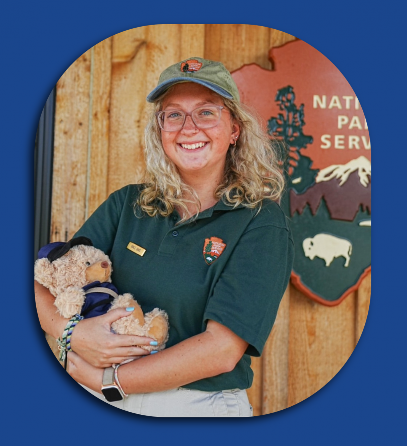 Woman stands in front of a National Park Service sign holding a stuffed bear.
